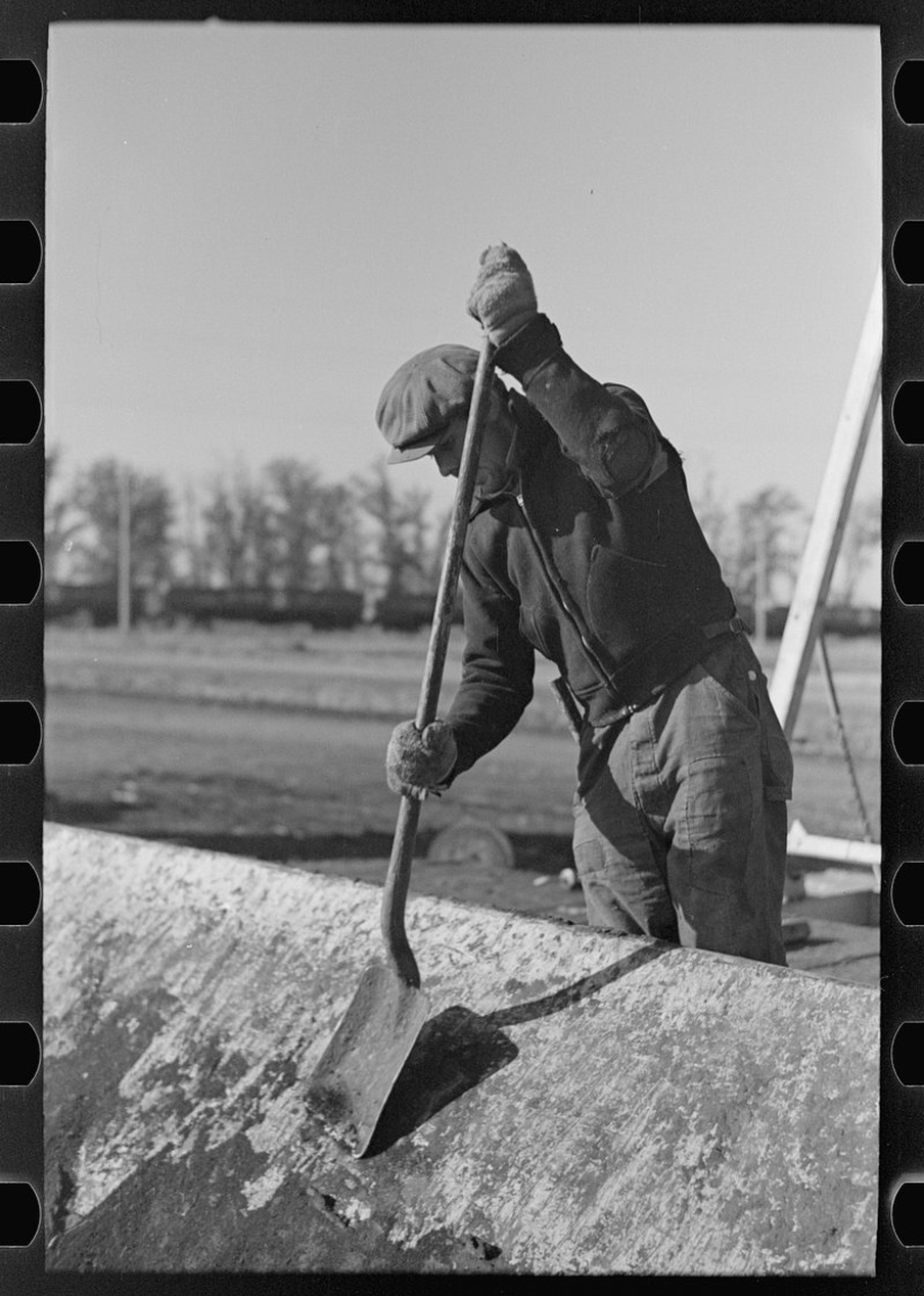 Vintage svartvitt fotografi som visar en person som använder ett långskaftat grävverktyg utomhus.